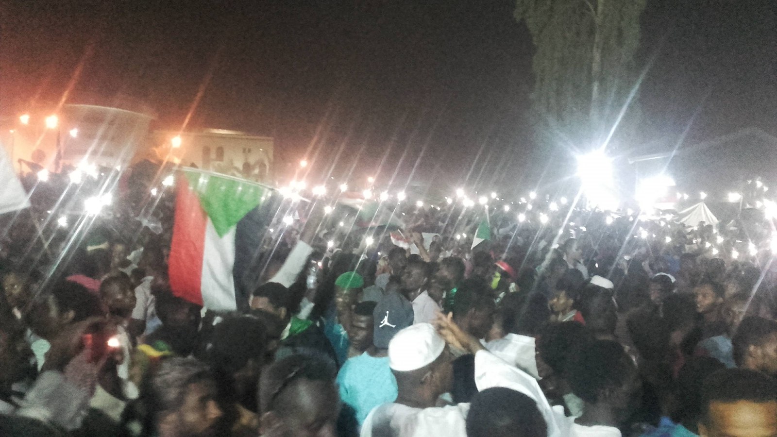 A nighttime public crowd under bright lights, with Sudanese flags raised above participants.