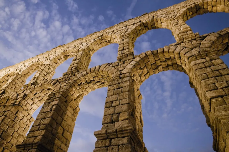 The stone arches of the Segovia aqueduct seen from below against a blue, clouded sky.